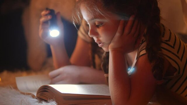 children in a wigwam tent play at night with a flashlight and read a book. happy family kid dream concept. children reading book with a flashlight dream. kid reading a book in a tent stay home house