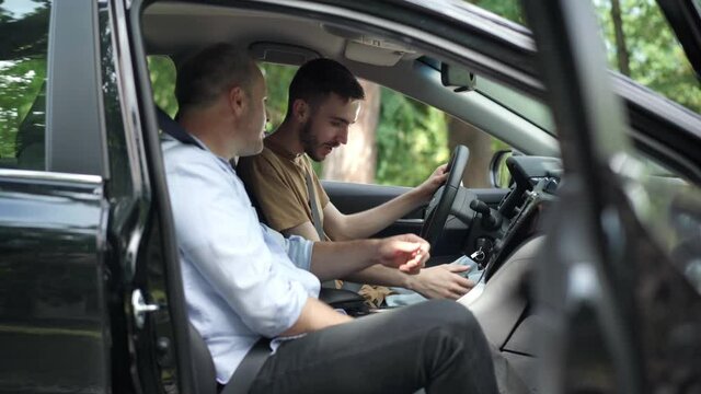 Side view of confident middle aged man teaching young cheerful guy driving. Positive excited Caucasian son or student learning as father or instructor talking. Lifestyle concept.