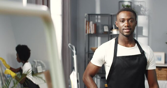 Portrait of young joyful handsome African American man in black apron standing in room looking at camera and smiling, female reflection in mirror cleaning furniture, clean up, housekeeping concept
