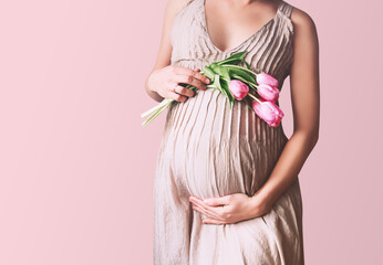 Beautiful pregnant woman with tulips flowers holds hands on belly. Young woman in dress waiting for baby birth. Pregnancy, Motherhood, Mother's Day Holiday concept.