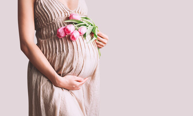 Beautiful pregnant woman with tulips flowers holds hands on belly. Young woman in dress waiting for baby birth. Pregnancy, Motherhood, Mother's Day Holiday concept.