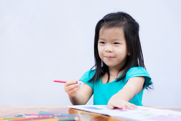 Cute girl sweet smile. Children are doing coloring activity with color pencil crayon on coloring book. On brown wooden table.Child enjoy making crafts. Isolated white wall background. Kid is 3-4 years