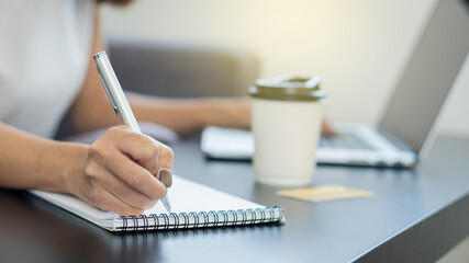 Hand of girl holding a pen in the notebook background with coffee mug and laptop