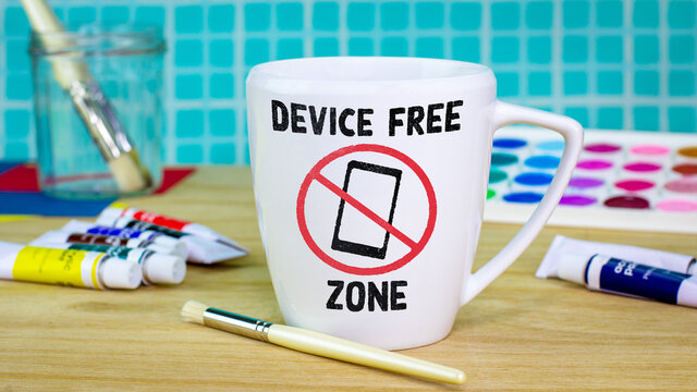 Device Free Zone mug with icon on mug on table surrounded by paints and brushes. Unplug from technology and plug into creativity for good mental health
