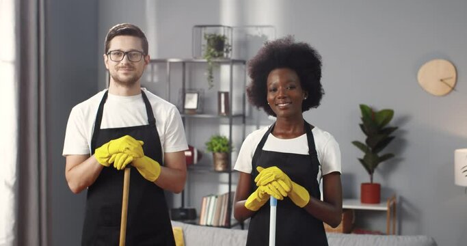Portrait of joyful positive mixed-races young team of professional cleaners in gloves and aprons looking at camera and smiling, multi-ethnic family couple clean up home, happy emotions