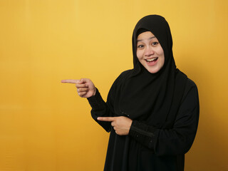 Asian muslim woman smiling and pointing to presenting something on her side, against yellow...