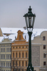 view of snow-covered statues and street lights on the old stones of Charles Bridge on the Vltava River and in the background the snow-covered roofs of buildings