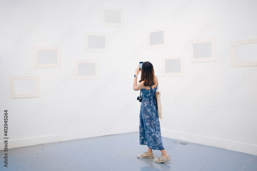 Wall mural Young asian thai woman in blue dress taking picture with smartphone in white gallery.