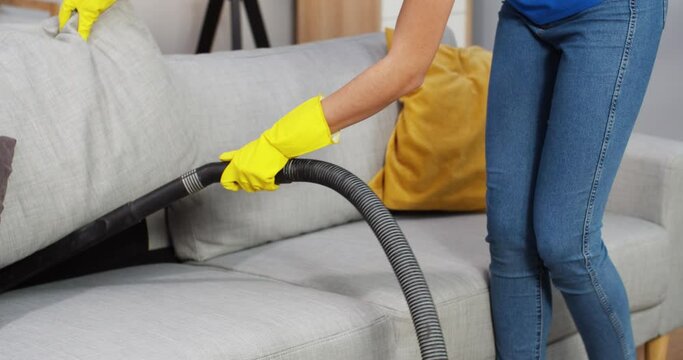 Close up shot of Caucasian female hands in yellow gloves using special vacuum cleaner to clean sofa, vacuuming couch in living room in house, cleaning furniture, clean up, domestic work concept
