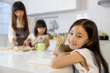 A cute little 7 years old Asian girl sitting and doing homework in kitchen while her mother...