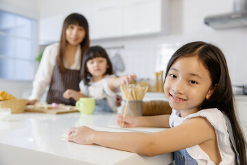 Obraz na płótnie Canvas A cute little 7 years old Asian girl sitting and doing homework in kitchen while her mother teaching her younger sister how to make food and cooking in blur background