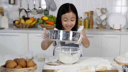 A little cute long black fair hair Asian girl, 7 years old, using riddle to sift flour to make bread and bakery with a curious and happy smile face in modern kitchen