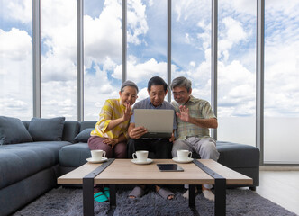 Fototapeta premium Three senior Asian people sitting together in living room and watching tablet computer screen and greeting for video call. Idea for learning and happy with new technology of older generation