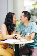 Happy young Chinese couple sitting at cafe table, touching hands and looking at each other