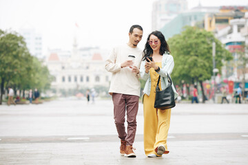 Happy young Chinese couple walking on city square and checking notification or map on smartphone