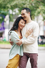 Beautiful happy young Chinese woman hugging boyfriend when they are standing outdoors