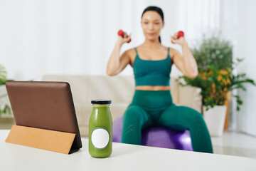 Fototapeta na wymiar Glass bottle of green smoothie on table next to tablet computer and fit woman exercising with dumbbells in background