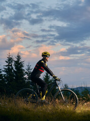 Young man cyclist riding bicycle on sunset in the mountains. Bicyclist wearing helmet, sports glasses and uniform. Side view. Evening sky on background. Concept of active lifestyle