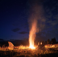 Beautiful view of night starry sky over grassy hill with camp tent, campfire and hikers. Young man and woman tourists having a rest near bonfire. Concept of hiking, night camping and relationship.