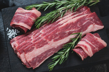 Close-up of raw marbled beef bacon with fresh rosemary on a black wooden chopping board, studio shot