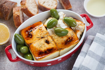 Red baking pan with blocks of feta cheese, giant green olives and bread over beige stone surface