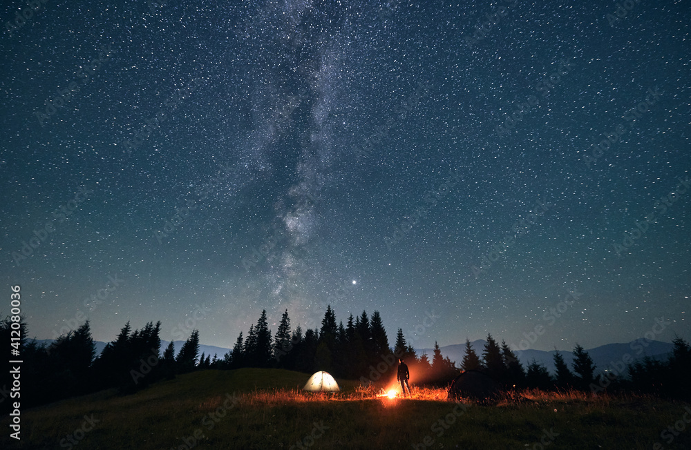 Wall mural silhouette of male traveler near illuminated camp tent under beautiful night sky with stars. magnifi