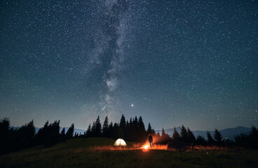 Silhouette of male traveler near illuminated camp tent under beautiful night sky with stars....