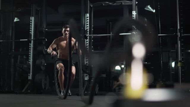 Athletic man performs exercises with battle ropes, endurance training in the gym, 4k slow motion.