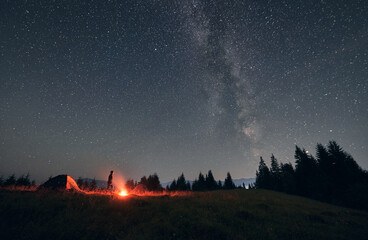 Male hiker standing near campfire and tent under beautiful night sky with stars. Magnificent view...