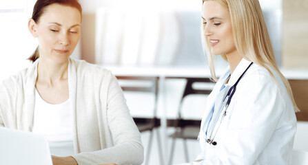 Woman-doctor at work in sunny hospital is happy to consult female patient. Blonde physician checks medical history record and exam results while using clipboard