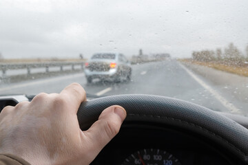 view of the driver's hand on the steering wheel of a car that is driving on the highway and in front of it an overtaking car makes a maneuver of changing lanes in rainy cloudy weather