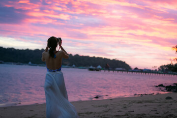 Young woman taking photos with her smartphone on beach during sunset or sunrise.