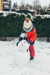Snow games, entertainment and fun. A cute Caucasian girl sculpts a snowman in the yard. Vertical orientation, authentic people