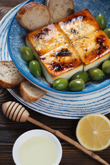 Blue bowl with baked feta cheese, green olives, baguette and olive oil, vertical shot on a brown...