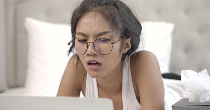 Young glasses asian woman lying in white bedroom. Girl sneezing to tissue paper while using laptop computer. Lady in casual clothes at house during quarantine. Social distance new normal concept.
