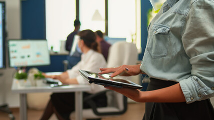Employee using tablet standing in office room while team of financial analysts working in background. Multiethnic coworkers respecting social distance in business company during coronavirus pandemic.