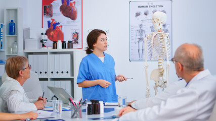 Medical young nurse presenting anatomy with human skeleton model describing the layout of the human spine. Explaining diagnosis to colleagues standing in front of desk in hospital meeting room.