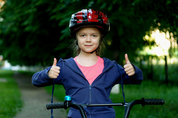 Naklejka premium Little girl learns to ride a bike in the park near the home. Kid shows the thumbs up on bicycle. Happy smiling child in helmet riding a cycling.