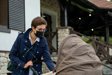 Beautiful woman with glasses and a medical mask walks with a baby in stroller. Quarantine,...