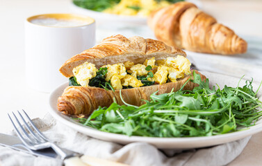 Delicious croissant sandwich with scrambled spinach eggs served with arugula on plate and coffee cup, light concrete background.