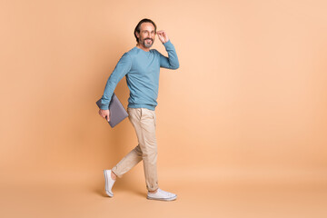 Fototapeta na wymiar Full length profile photo of man walk hold laptop wear specs blue sweater pants sneakers isolated beige color background