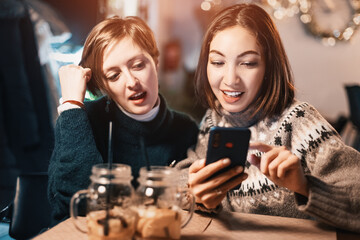 Two happy women sit in a cafe, chat with each other and watch the news on social networks on their mobile phone. The concept of relationships and communication