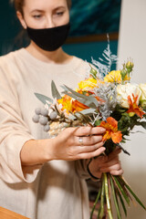 Florist works with colors. Flower seller chooses flowers for future bouquet. Flowers shop worker in a mask standing in flower shop and checking flowers in glass vase.