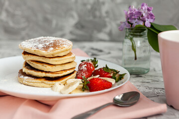 A stack of pancakes with a pink tea mug and  fresh strawberries on a white plate on a light background. A small bouquet of lilacs in a glass vase stands next to the Breakfast.