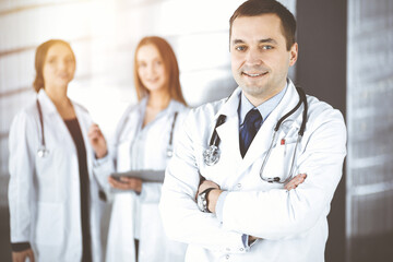 Professional middle aged doctor with a stethoscope and crossed arms at work, standing together with his female colleagues in a sunny clinic. Perfect medical service in a hospital. Medicine and