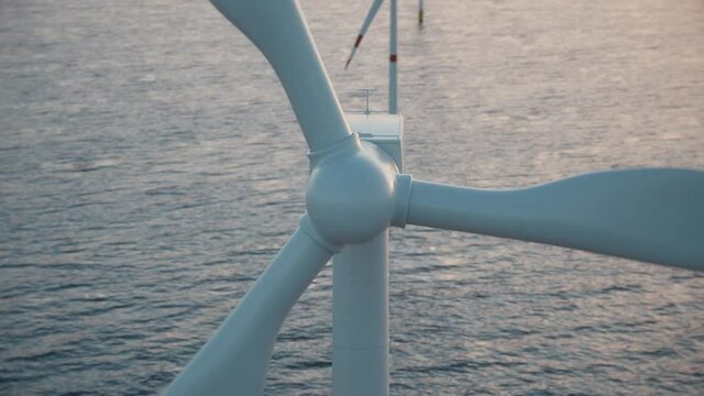 Camera moves around the head of a wind turbine to reveal an aerial view of a large wind farm in the sea against a low sun. Green and renewable energy concept. Realistic high quality 3d animation. 