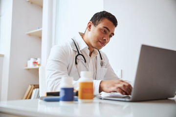 Fototapeta premium handsome confident man therapist in white robe with stethoscope sitting at the work desk in cabinet