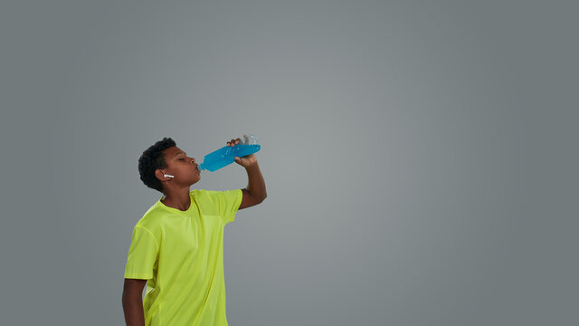 Dehydration During Physical Activities. Side View Of A Sporty Teenage African Boy Drinking Blue Energy Drink While Standing Against Grey Background