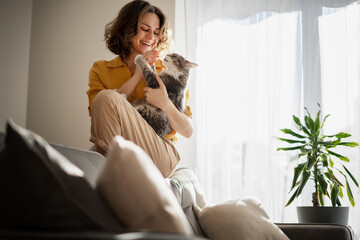Beautiful cheerful young woman with a cute gray cat in her arms at home in the interior, friendship...