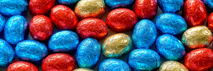 Closeup of many chocolate easter eggs wrapped in blue, red and golden foil. Easter banner background.